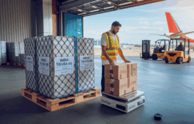 Ground staff loading pallets and parcels for India-to-USA air freight with labeled cargo showing per-kg shipment details at an Indian airport.